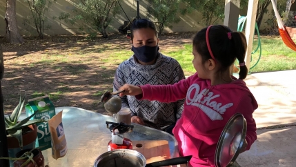 A girl wearing a pink shirt drinks a beverage with her mother, wearing a grey shirt, outdoors at a table. 