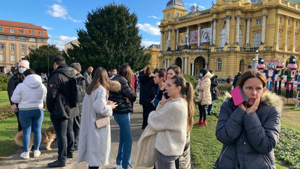 A crowd of people are shown standing outside, many holding cell phones, with classical European buildings in the distance.