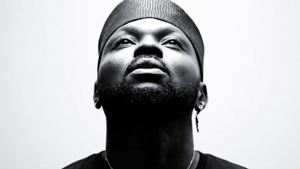 A black and white photograph of musician Shirazee wearing a traditional hat and looking up.