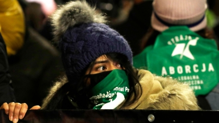 A crowd of people are shown with a woman in the center holding a banner and wearing a winter hat and green scarf with writing on it.
