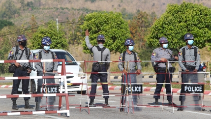 A line of six security authorities are shown standing next to a metal baracade blocking a road with one man holding his hand up stopping an approaching car.