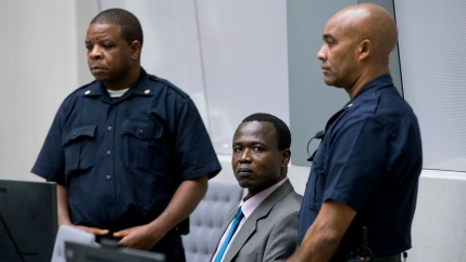 Dominic Ongwen is shown sitting and wearing a gray suit while two security officers are shown standing on either side of him.