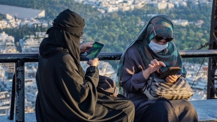 Two women sit outside, using their cell phones. They are wearing face masks in addition to head coverings. 