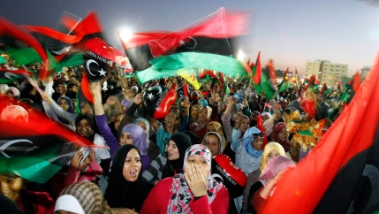 A large crowd of people are shown with many carrying the Libyan flag in a blurred motion photograph.