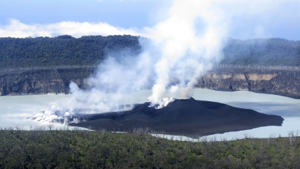 A dark volcano with smoke rising out of it surrounded by a body of water. 