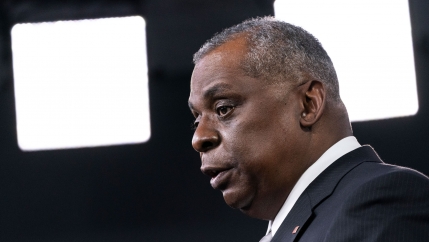 Secretary of Defense Lloyd Austin is shown in a close-up photograph from the side with bright white lights in the distance.