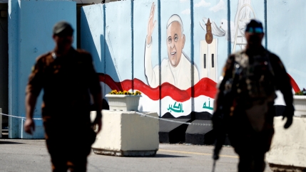 Two police officers are show in shadow in the nearground with a picture of Pope Francis on a wall in the background with the Iraqi flag painted at the bottom.