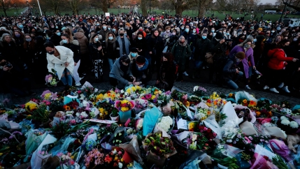 A large crowd of people are shown standing vigil next to hundreds of flowers laid on the ground in memory of Sarah Everard.