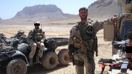 A soldier in uniform stands next to a man in a military vehicle with rugged mountains in the background.