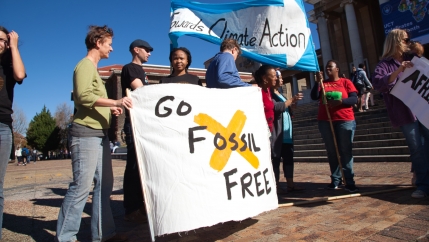 A group of people hold up a large white sign with black letters and a yellow X through it. 