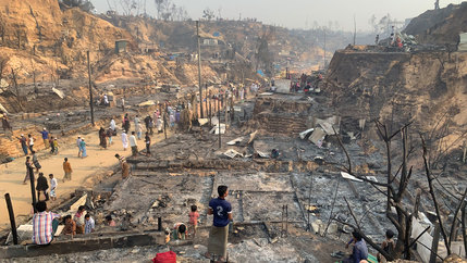 Several people are shown standing in the burned down ruins of the Balukhali refugee camp.