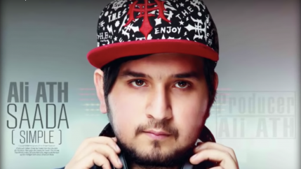 Ali ATH, rapper wearing a red and black cap and holding headphones around his neck. 