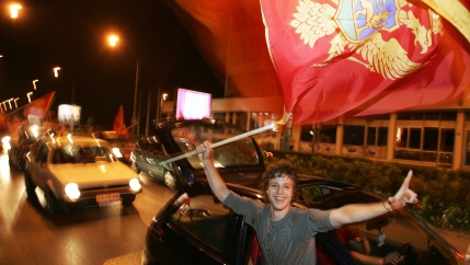 A man holds a flag and sticks his arm out of a car window, smiling and celebrating. 