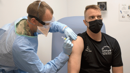 Army captain in scrubs administers COVID-19 vaccine to man in black shirt
