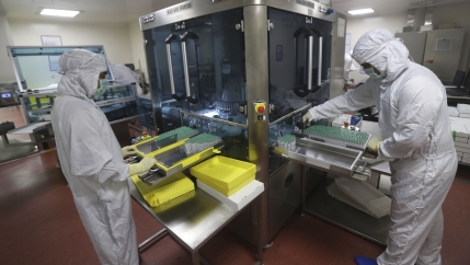 Two lab technicians dressed in white protective gear organize vials. 