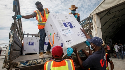 Men in orange and yellow jackets lift a box of vaccines from COVAX off of a truck bed
