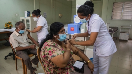 A man and woman are shown sitting in chairs while medical workers administer vaccinations.