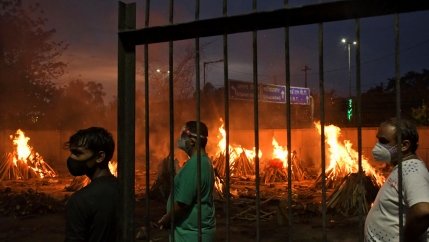Three men are shown standing and wearing protective face masks with several funeral pyres flaming in the distance.