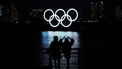 Two people are shown standing at a railing next to a body of water where the Olympic rings are in the destance illuminated in white.