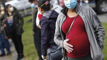 A pregnant person wearing a red shirt holds her belly, wearing a face mask and gloves. 
