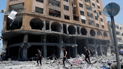 Three men are shown walking in the street covered in rubble and past a bombed building with the bottom floor blown out.