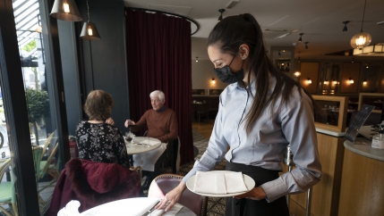 A waiter wearing a mask and an apron with her hair pulled back tends to a dining table. 