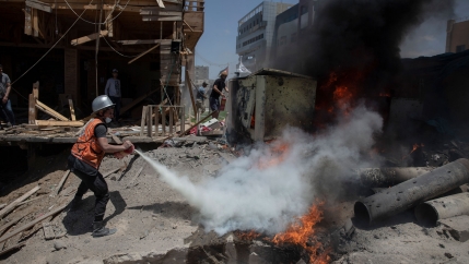 A man is shown wearing an orange safety vest and silver helmet and spraying a fire working to extinguish it.