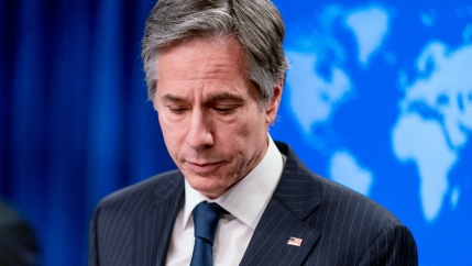 Secretary of State Antony Blinken is shown wearing a blue suit and tie with a US flag pin while looking down.