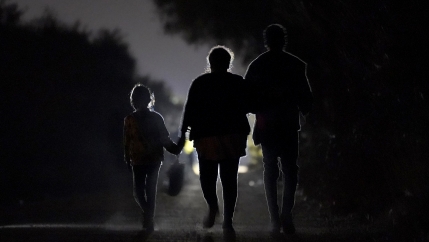 Figures of a 7-year-old migrant girl walking with a woman and unidentified man silhouetted at night