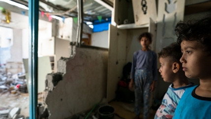 Three young children are shown looking off to a room damaged by rocket attack.