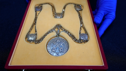 Person with blue gloves presents a large medal in a yellow and red box