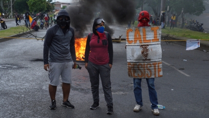 Protesters block a road in downtown Cali using ropes and burning wood. One of them uses a home made shield that says “Cali Resists