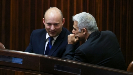 Yamina party leader Naftali Bennett wearing a dark suit and sitting while speaking with Yesh Atid party leader Yair Lapid on his left.