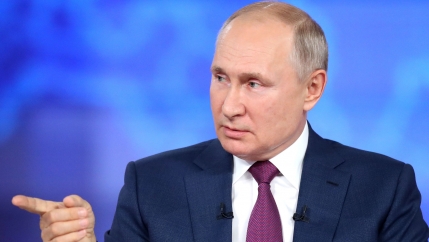 Russian President Vladimir Putin is shown wearing a blue suit and purple tie while pointing with his right hand.