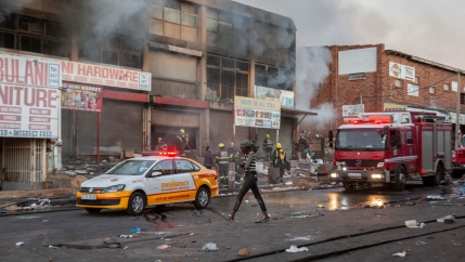 A woman is shown walking past a row of smoking buildings from a fire and fire trucks.