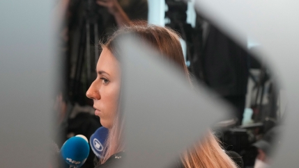 Belarusian Olympic sprinter Krystsina Tsimanouskaya is shown through a frosted glass window speaking into microphones.