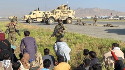 Several US military personnel are show standing guard next to the runways of the Kabul airport with two large military vehicles in the distance.