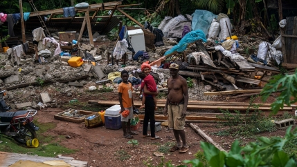 A man is shown without a shirt on standing next to a woman holding a small child with the rubble of their home in the background.