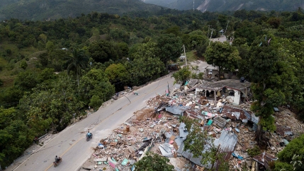 Several buildings are shown reduced to rubble on a road with two people passing by on motorcycles.