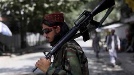 A bearded man is shown in profile wearing sunglasses, fatigues and carrying a large gun over his shoulder.