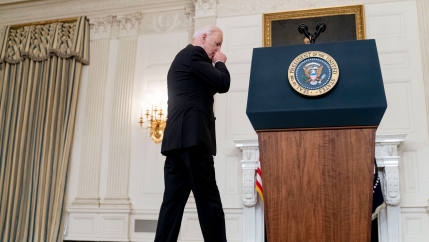 US President Joe Biden is shown wearing a dark suit and walking toward a podium with the President of the United States seal on it.