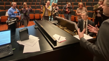 Group of people stand around a pianist in an auditorium