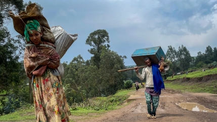 A women is shown in the nearground carrying large sacks over her back and walking with a man in the distance carrying a large blue crate.
