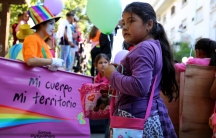 A young girl holds a balloon. Behind her are protestors carrying signs and children with more balloons. One person is dressed as a clown with a sign that says,