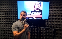 Bassem Youssef stands in front of a microphone in a recording studio. Behind him is a screen lit up with the title of his new podcast: