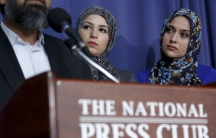Two women wearing head scarves watch a man at a podium.