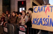 A crowd stands behind a metal gate. One holds a sign that says