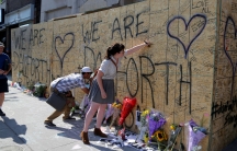 People leave messages, flowers and other trinkets on a plywood wall that says