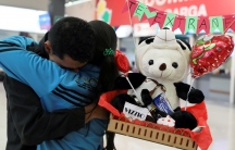 Two people embrace in a bus station. One of them holds a greeting balloon, candy and a stuffed animal.