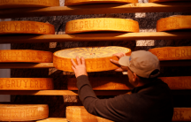 a man stands in front of a wall full of wheels of cheese